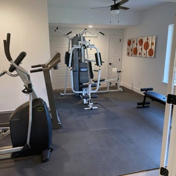 A clean, home gym with gym flooring covering the floor. The room contains an elliptical, a treadmill, and a multi-station weight machine