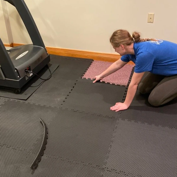 A woman in a blue t-shirt and brown leggings installs gym flooring in a home gym.